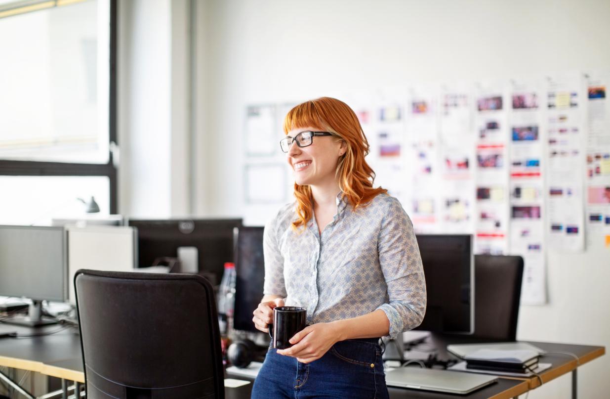 A woman in an office wearing glasses and holding a coffee cup smiling