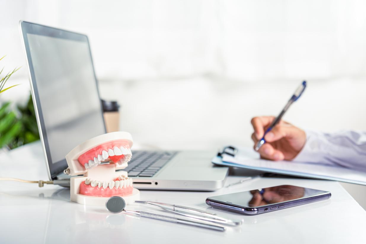 Someone with a laptop open and writing on a clipboard with a model of teeth and dental tools on the table