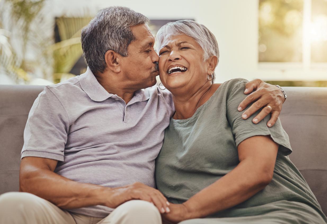 Happy senior couple sitting next to each other with husband's arm around wife while he kisses her on the cheek.