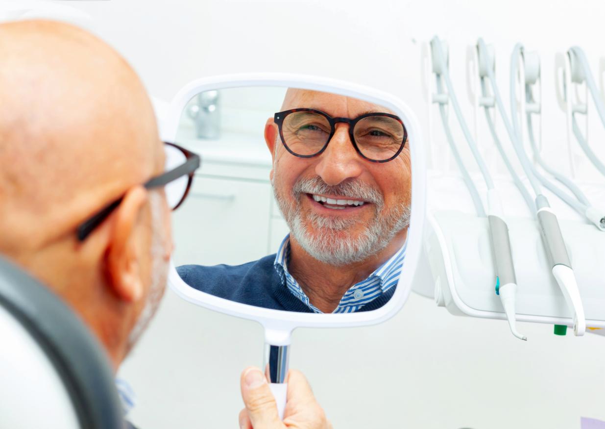 Happy senior man at dentist viewing his beautiful smile in the reflection of a mirror.