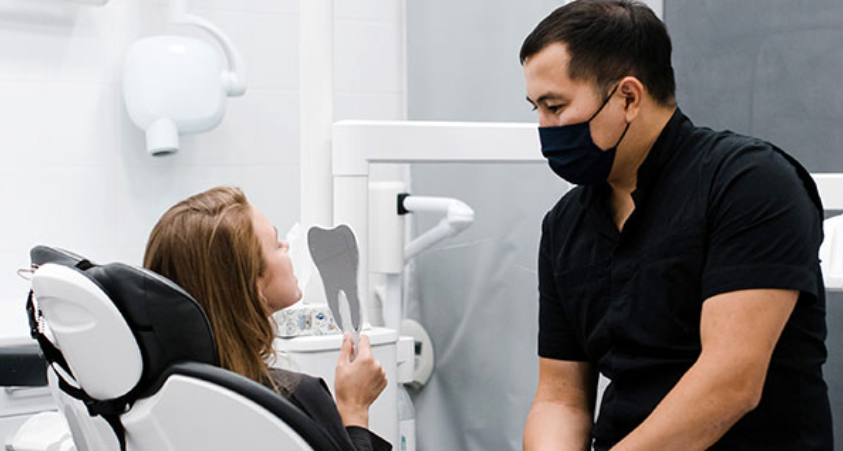 a dentist sits next to a patient who looks at her teeth in a mirror