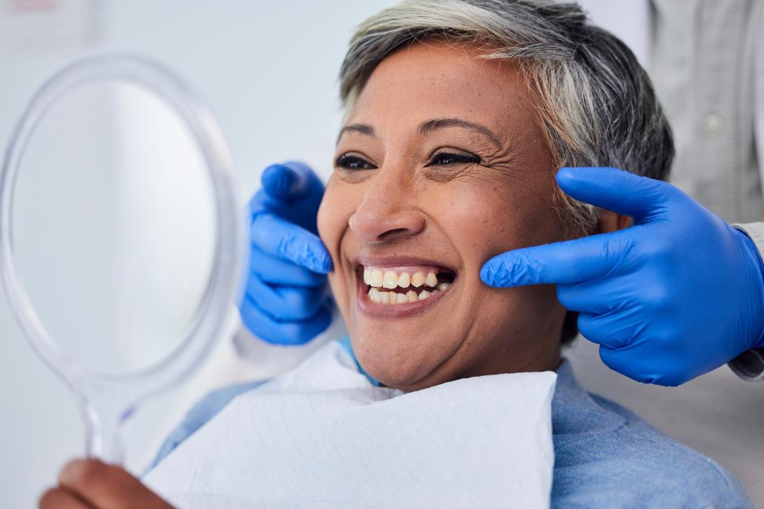 Senior woman dental patient smiling while looking at reflection in mirror and with dentist's hands gently on her face.
